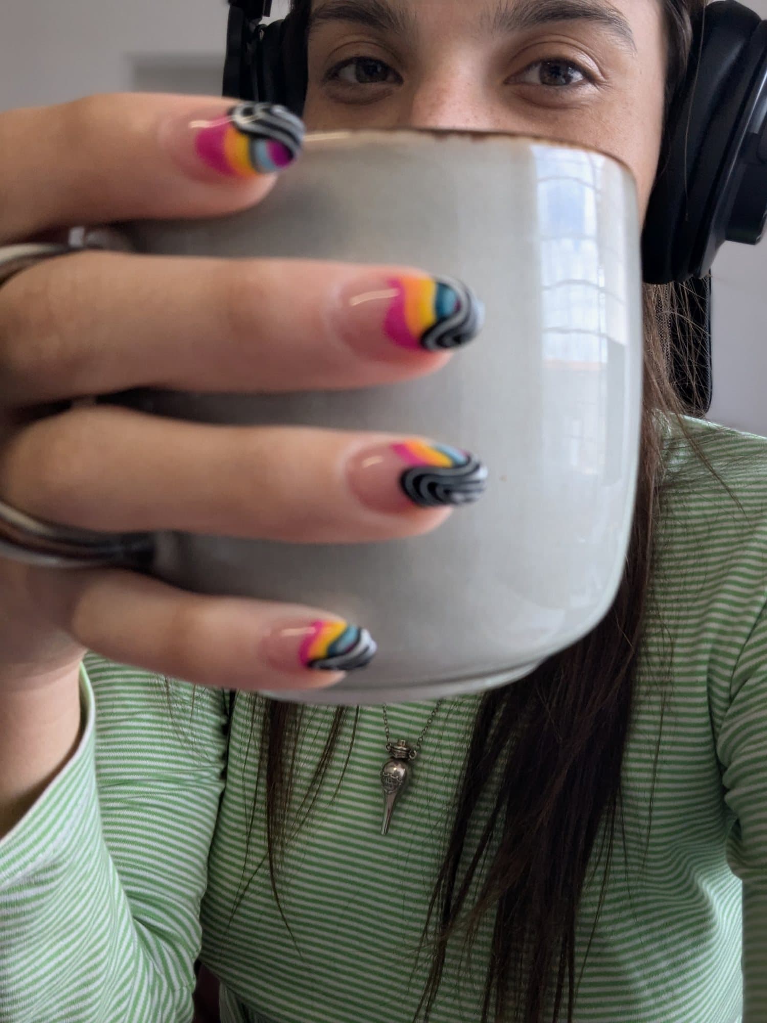 Val tomando de una taza grande, con audífonos y uñas de colores