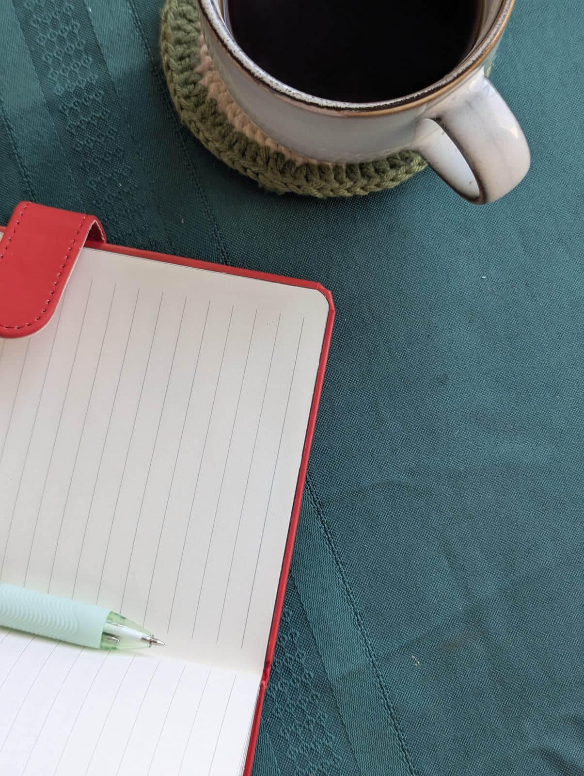Cuaderno rojo abierto, un bolígrafo y una taza de café sobre un mantel verde